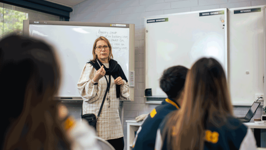 A teacher stands at the front of a class, talking. 